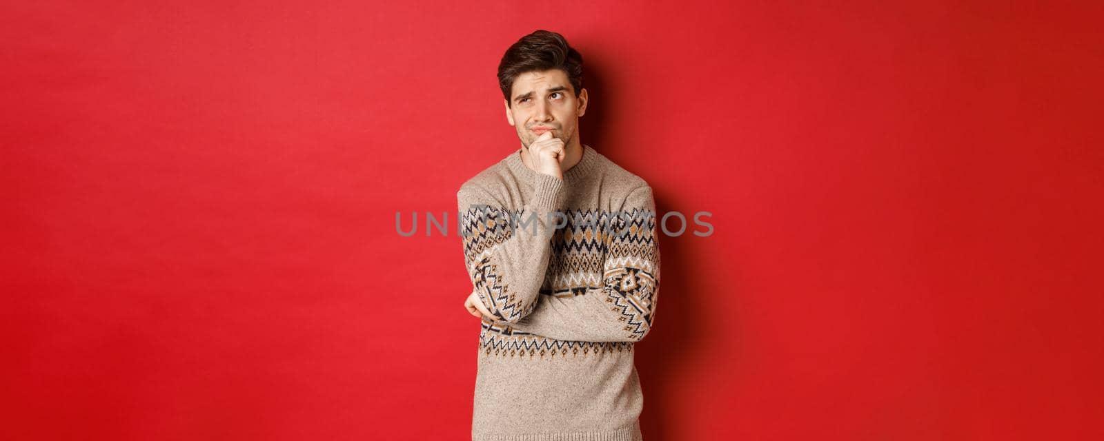 Portrait of confused man in winter sweater, thinking about christmas presents, looking troubled, searching for good gifts, standing over red background by Benzoix