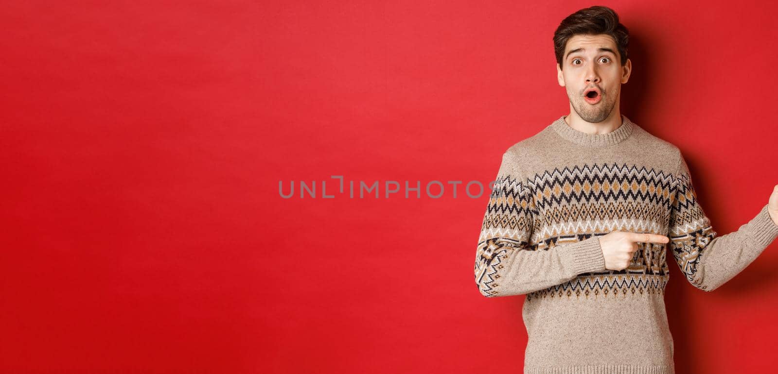 Concept of christmas celebration, winter holidays and lifestyle. Amazed handsome guy in xmas sweater pointing fingers right at logo, showing advertisement, standing over red background by Benzoix