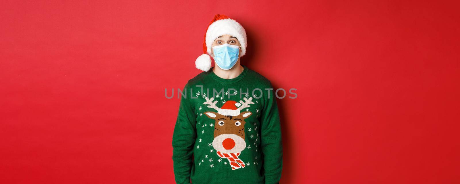 Concept of new year, covid-19 and social distancing. Surprised guy in santa hat, medical mask and christmas sweater looking amazed at camera, standing over red background.