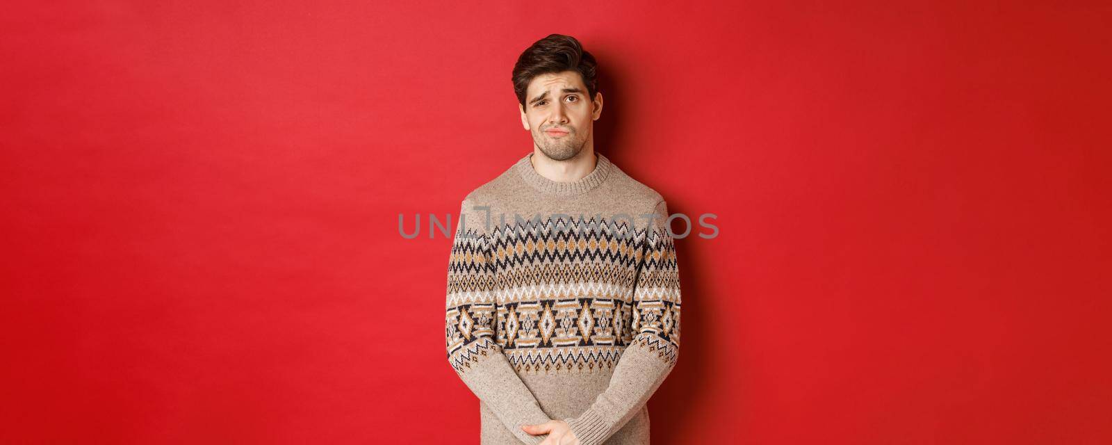 Portrait of gloomy and sad caucasian man in christmas sweater, frowning and pouting indecisive, having bad new year, standing over red background.