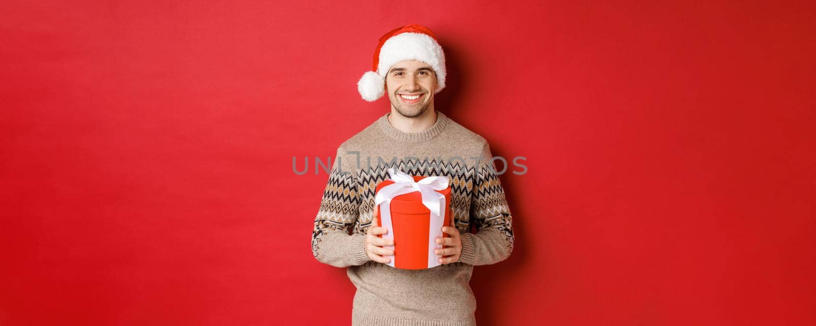 Portrait of handsome man holding a present, wishing happy holiday, standing in santa hat and christmas sweater against red background by Benzoix