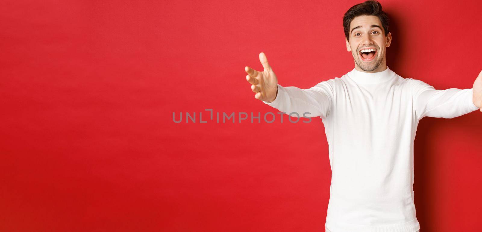 Image of happy, friendly man in white sweater, reaching hands and smiling while greeting guests to christmas party, waiting for hug, standing over red background.