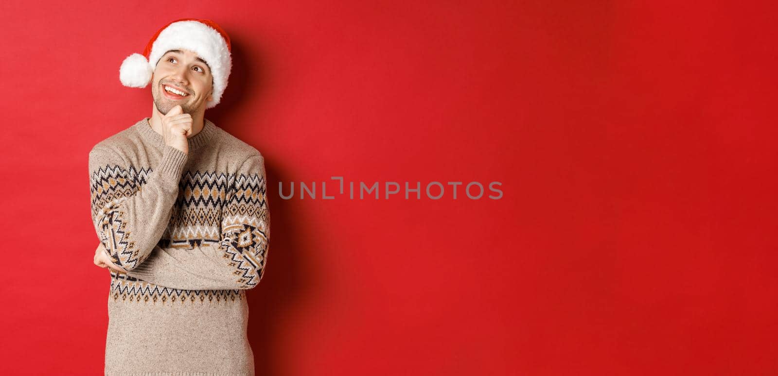 Image of happy young man in santa hat and christmas sweater, imaging something, thinking about new year gifts and smiling, looking at upper left corner, standing over red background by Benzoix