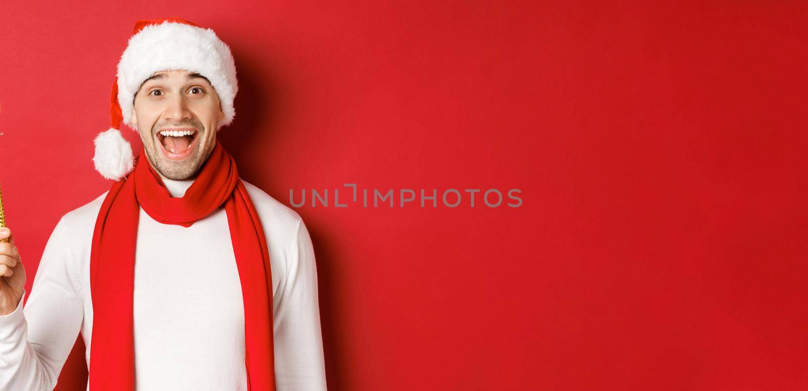 Concept of christmas, winter holidays and celebration. Handsome man celebrating new year and having fun, holding sparkler and smiling, wearing santa hat, standing over red background.