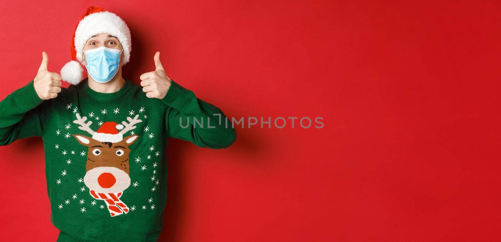 Concept of new year, covid-19 and social distancing. Cheerful handsome man in santa hat and medical mask, showing thumbs-up, enjoying christmas party, standing over red background.
