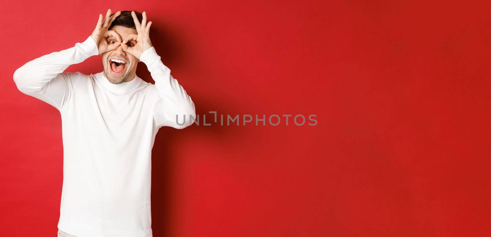 Portrait of handsome guy in white sweater, making funny mask with fingers, looking happy and smiling, standing over red background.