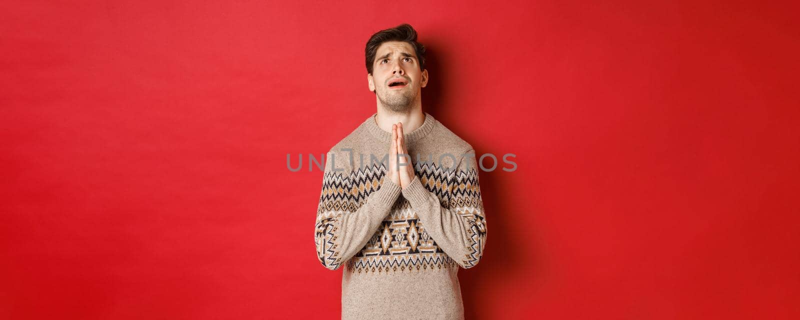 Image of nervous and hopeful man praying to God, begging for help with christmas, wearing winter sweater, looking up and pleading, standing over red background.