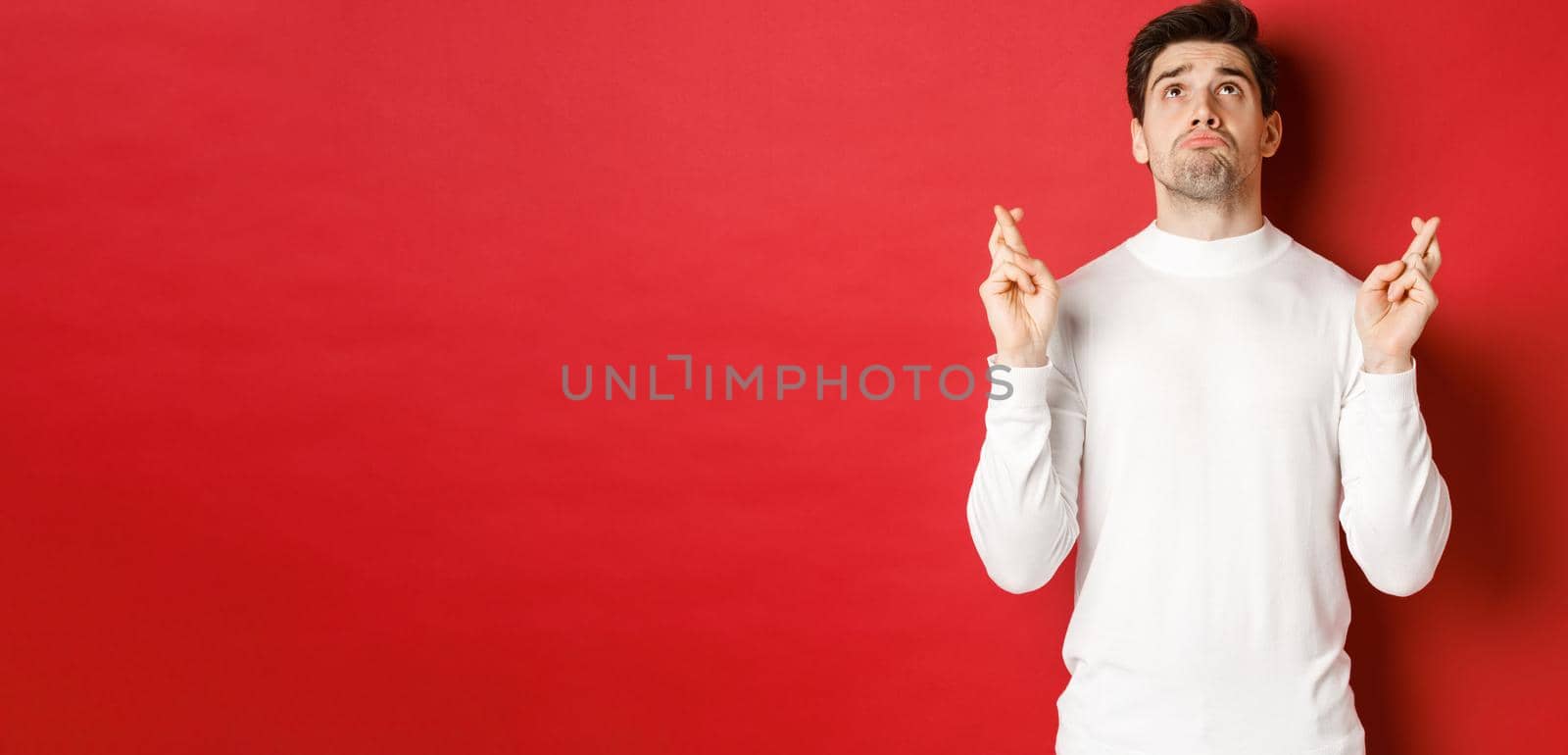 Portrait of sad and hopeful man making a wish, begging god and looking up, crossing fingers for good luck, standing over red background.