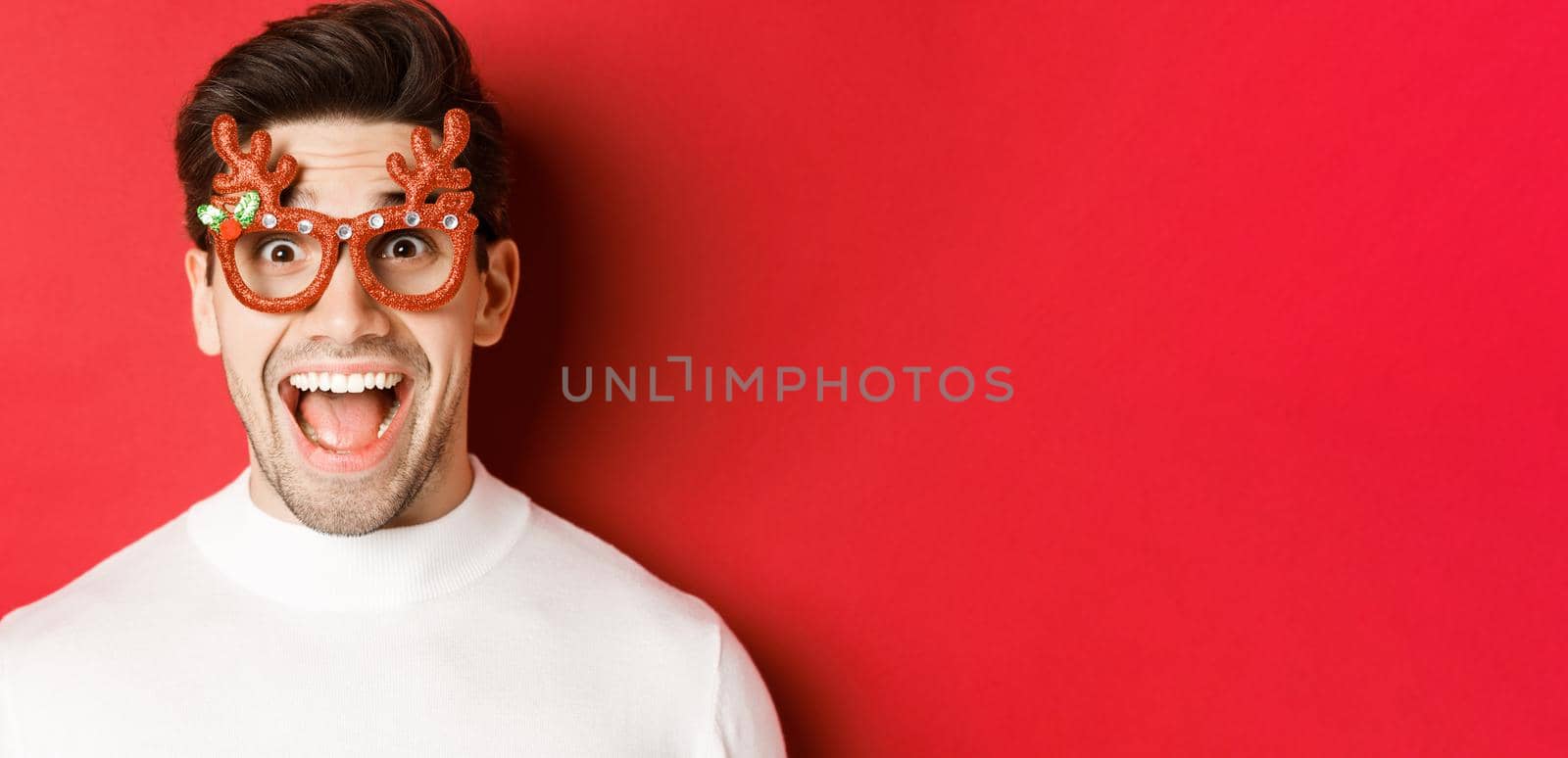 Concept of winter holidays, christmas and celebration. Close-up of attractive guy with bristle, wearing new year party glasses and looking amazed at promo offer, standing over red background by Benzoix