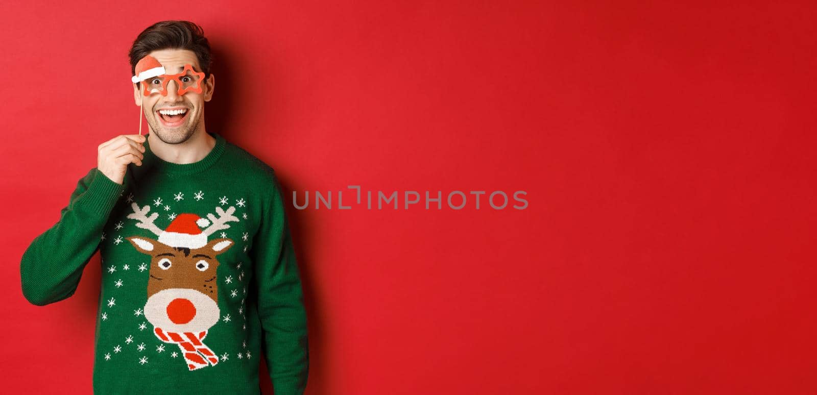 Portrait of handsome smiling man in christmas sweater and party glasses, looking surprised and happy, celebrating new year and having fun, standing against red background.
