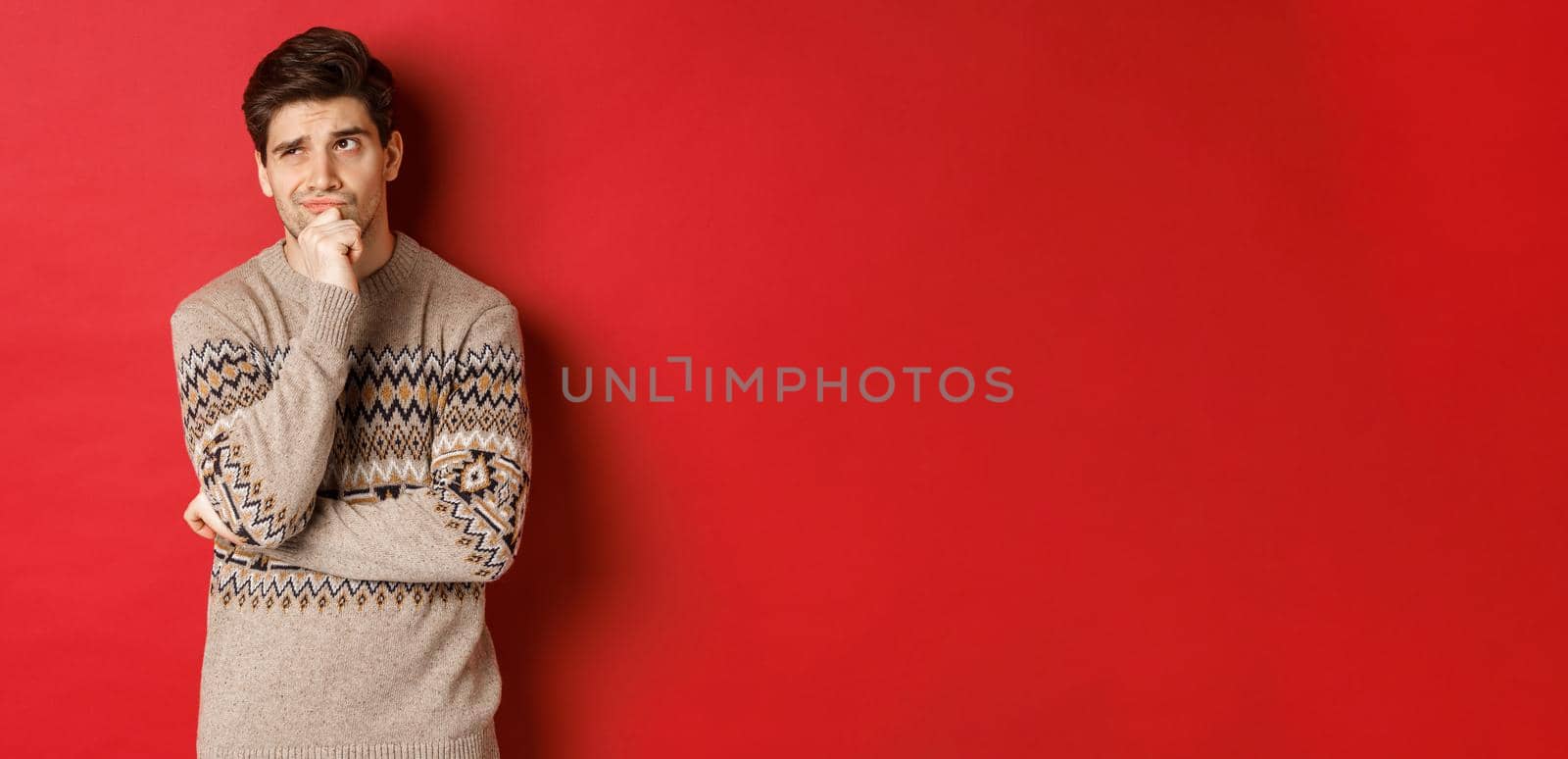 Portrait of confused man in winter sweater, thinking about christmas presents, looking troubled, searching for good gifts, standing over red background.