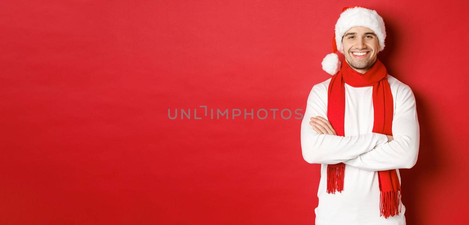 Handsome caucasian guy with bristle, wearing santa hat, scarf and white sweater, cross arms on chest and smiling happy, standing against red background.