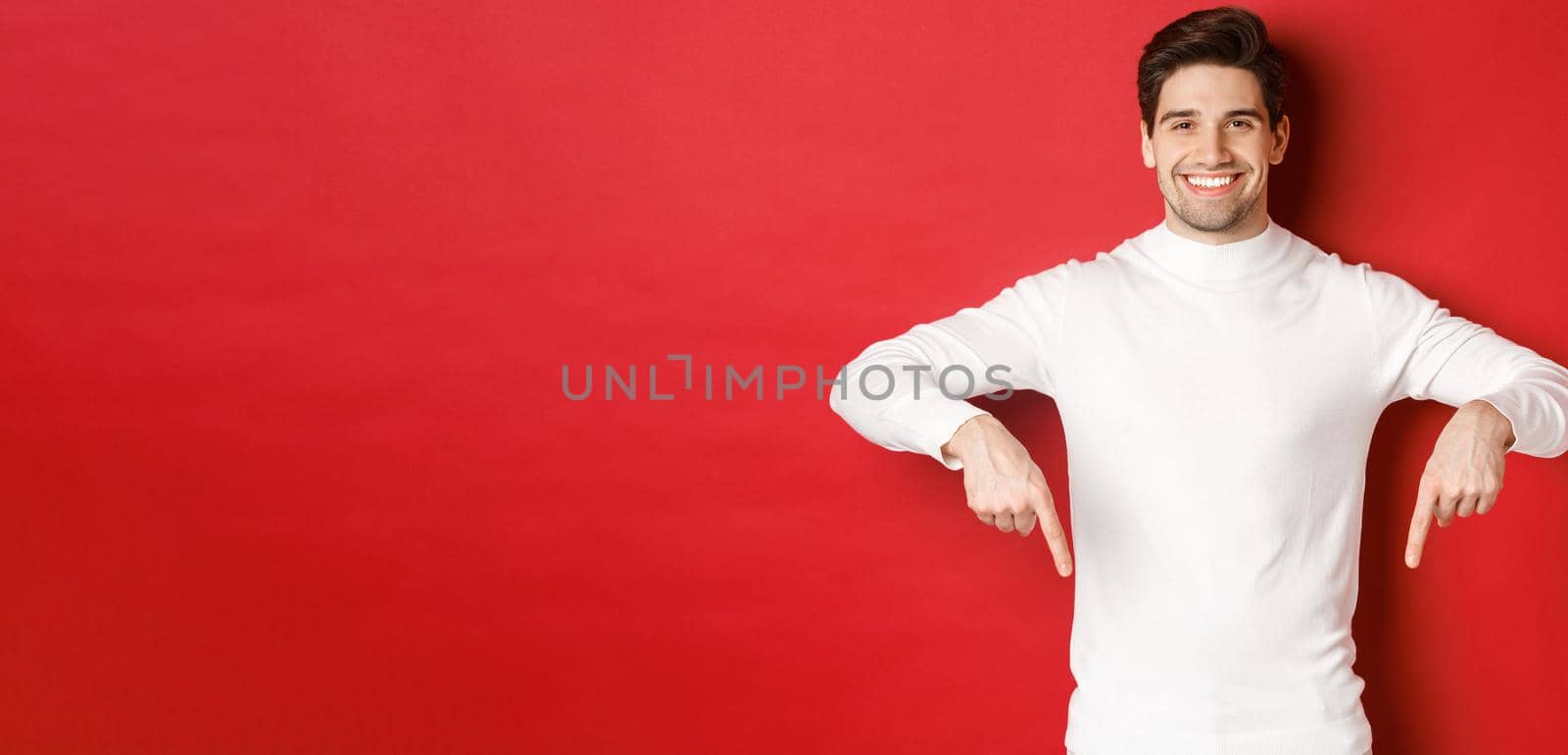 Image of smiling handsome man in white sweater inviting visit page, pointing fingers down and showing christmas banner, standing against red background by Benzoix