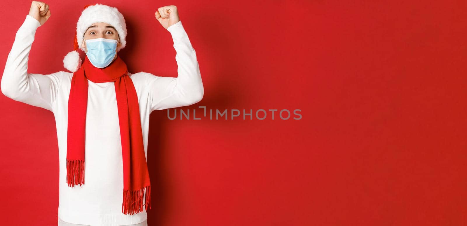 Concept of covid-19, christmas and holidays during pandemic. Portrait of happy man in santa hat and medical mask, rejoicing and celebrating new year, standing over red background.