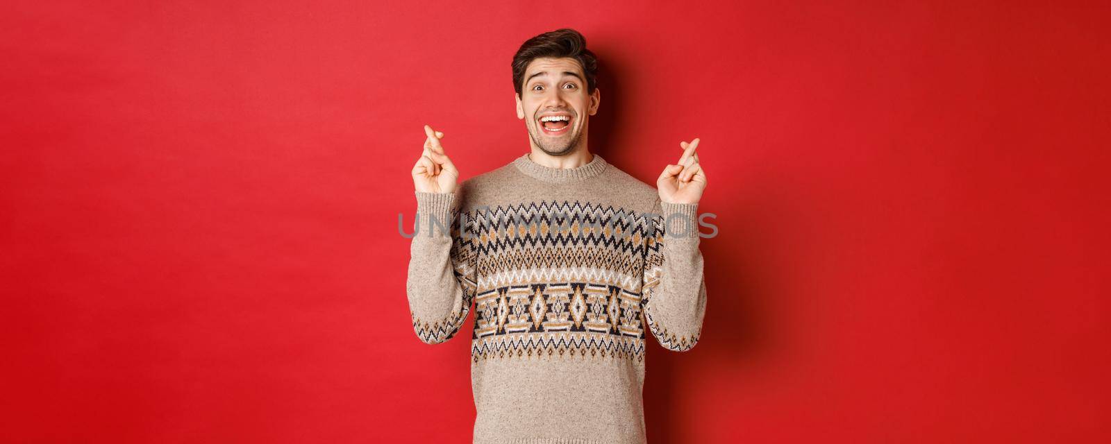 Image of excited and hopeful young man making wish, wearing winter sweater, crossing fingers for good luck and smiling, anticipating christmas eve, standing over red background by Benzoix