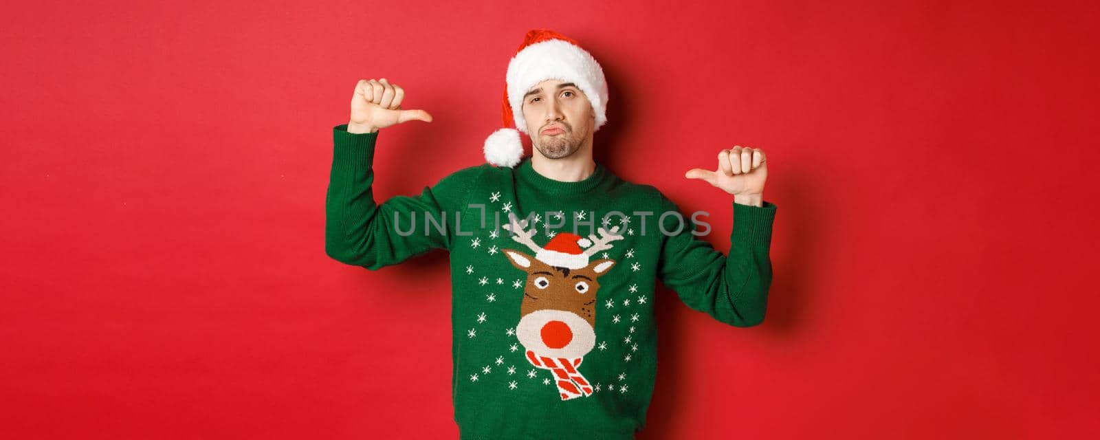 Image of handsome and confident young man in green sweater and santa hat, pointing at himself, celebrating christmas, standing over red background.