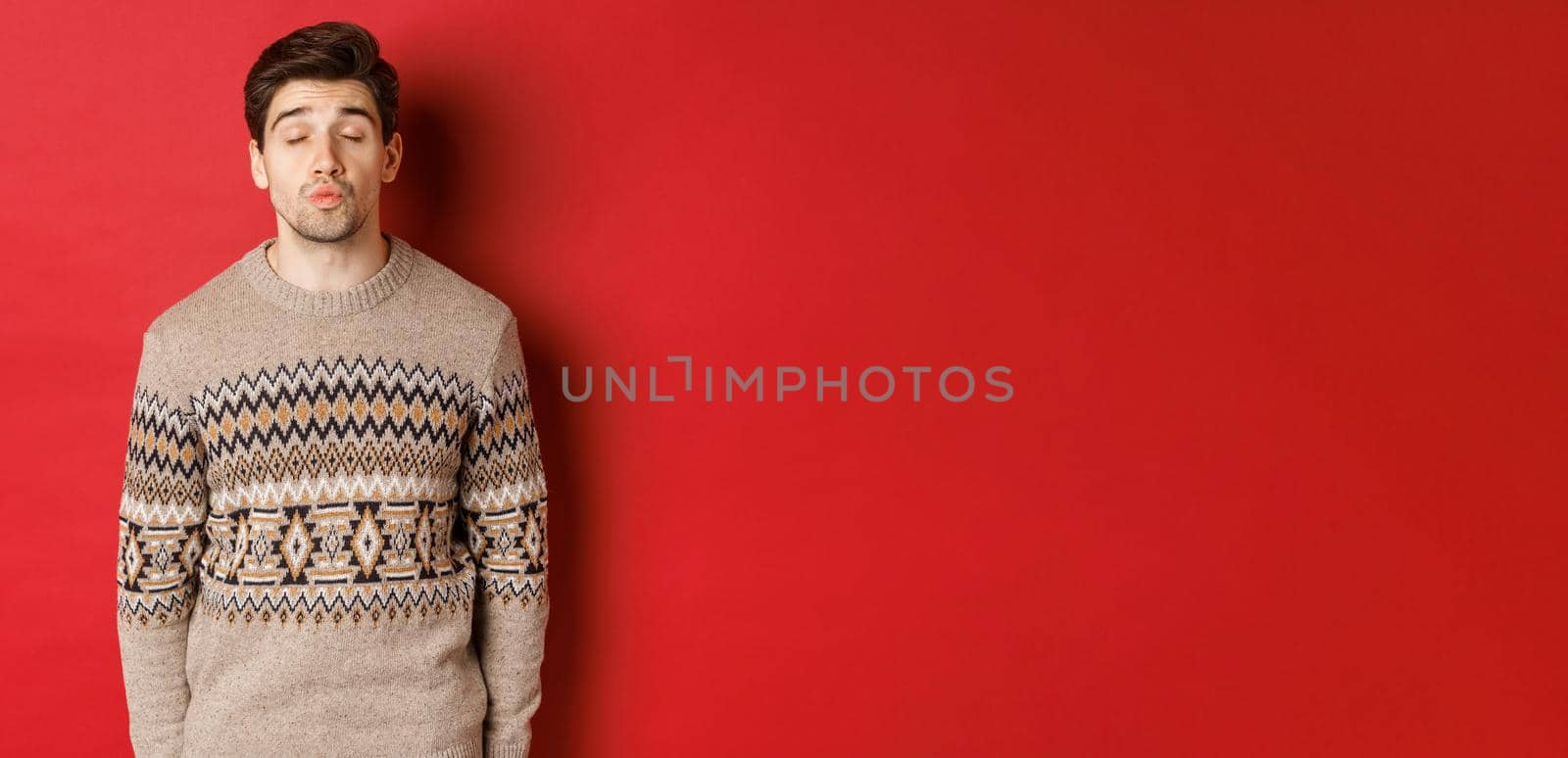 Image of handsome man in christmas sweater pucker lips and close eyes, waiting for kiss under mistletoe, standing over red background by Benzoix