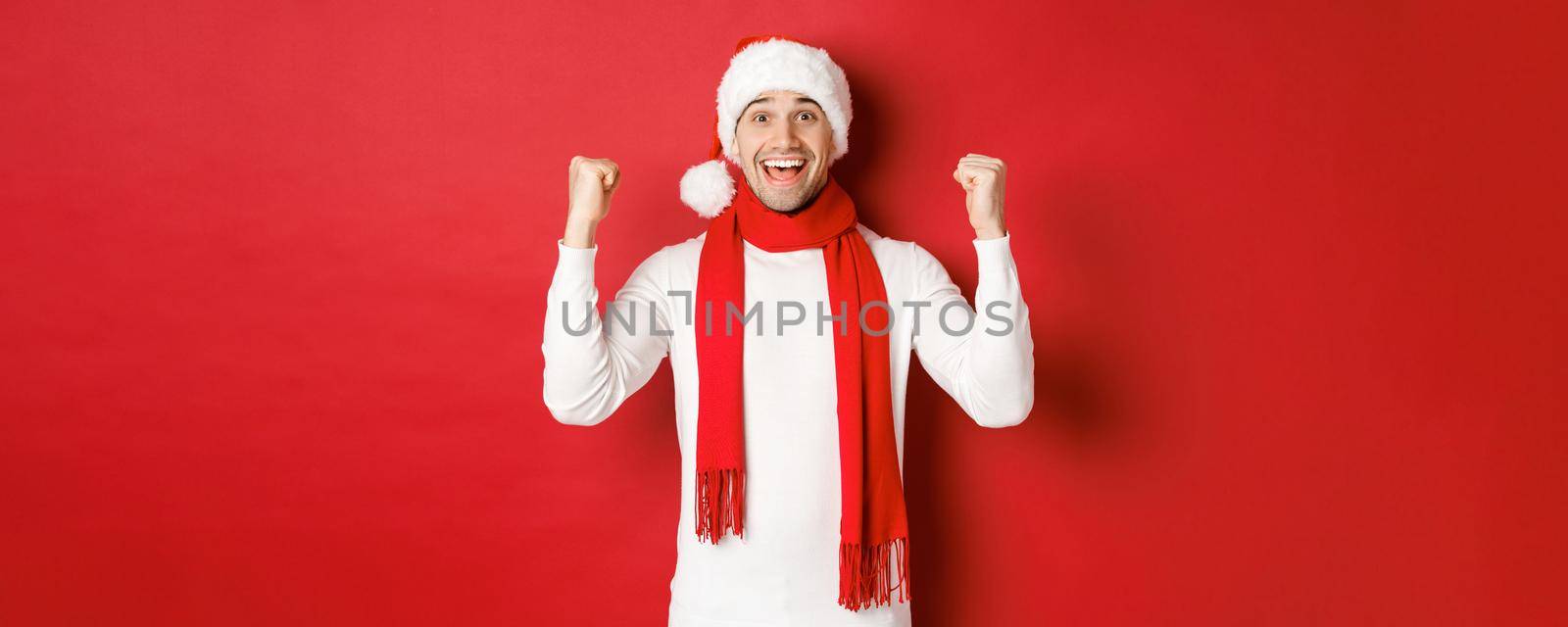 Portrait of happy and excited man in santa hat and scarf, rejoicing and winning something, celebrating new year, standing over red background.