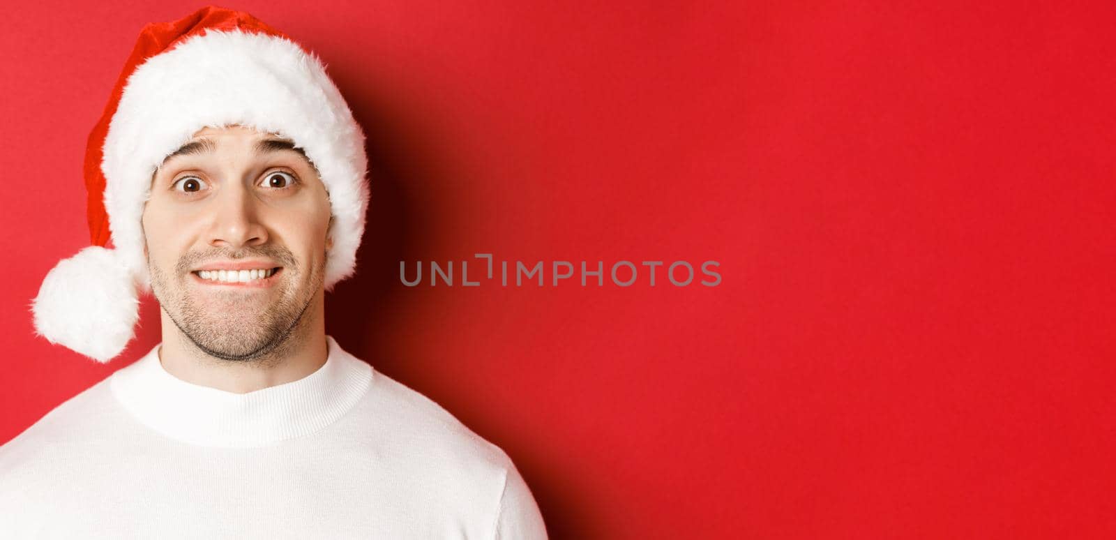 Close-up of handsome guy in santa hat, biting lip and looking with temptation at something he wants, standing over red background.