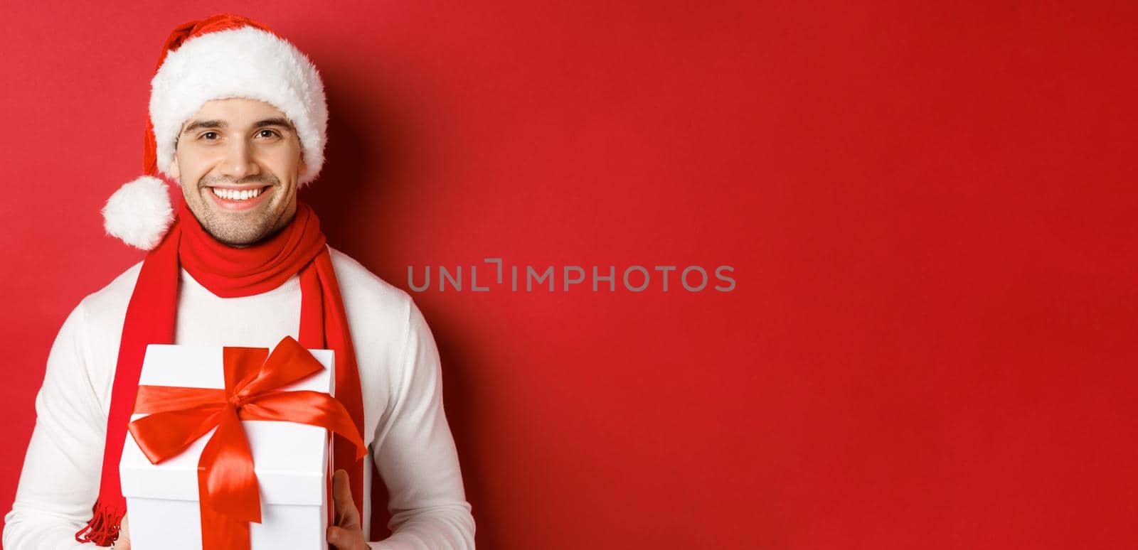 Concept of winter holidays, christmas and lifestyle. Close-up of attractive bearded guy in santa hat and scarf, holding a present, giving new year gift and smiling, red background by Benzoix