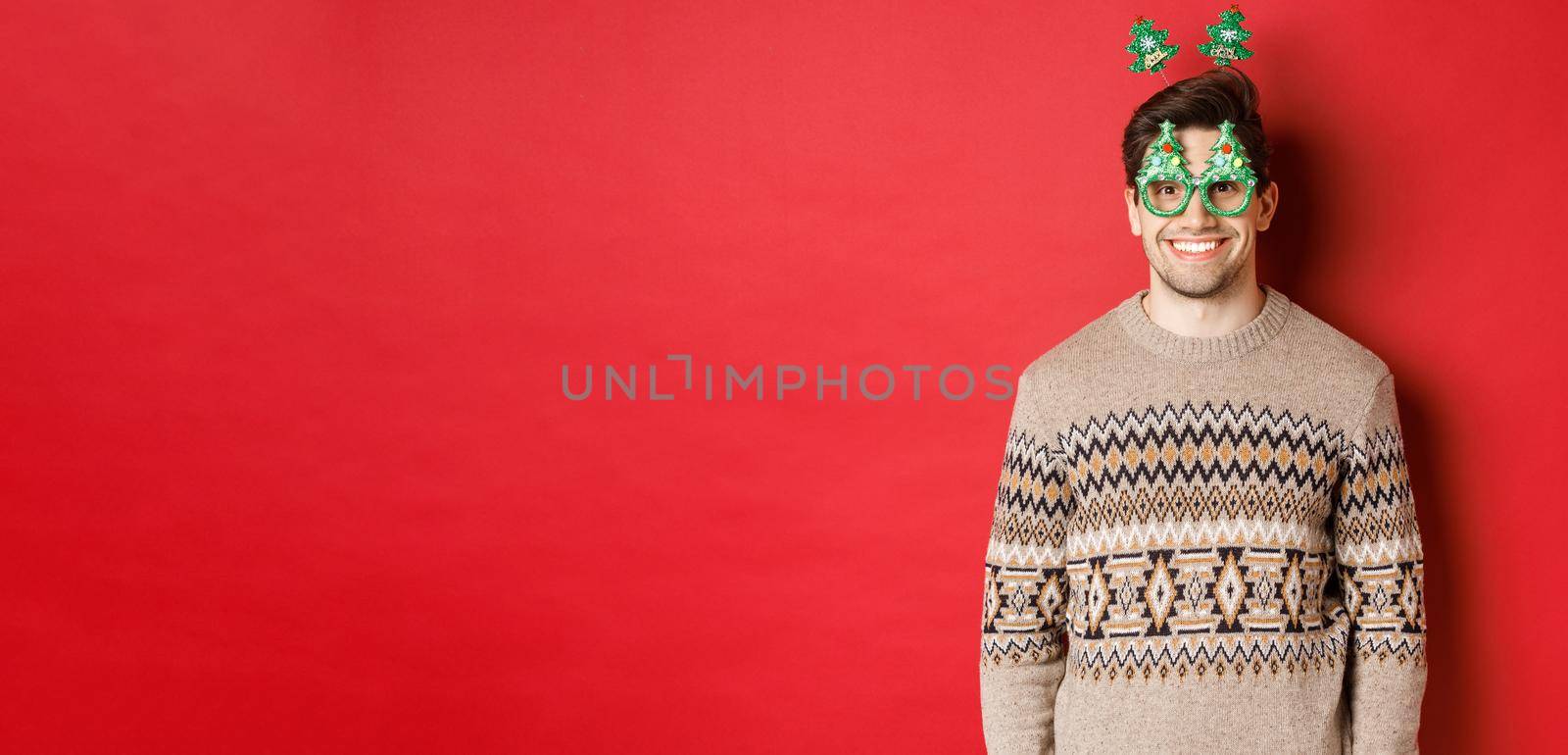 Portrait of happy and cute man in winter sweater and party glasses, celebrating new year or christmas, smiling while standing over red background.