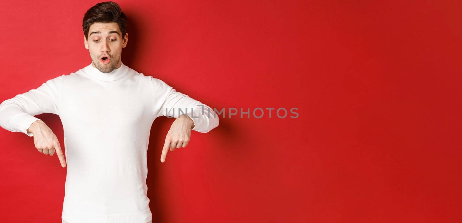 Portrait of surprised good-looking guy in white sweater, looking and pointing fingers down at logo, standing against red background.