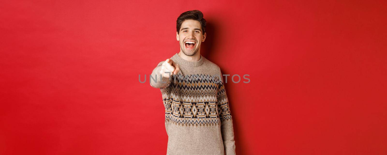Concept of christmas celebration, winter and lifestyle. Happy attractive man in xmas sweater, wishing happy holidays and pointing at camera, standing over red background by Benzoix