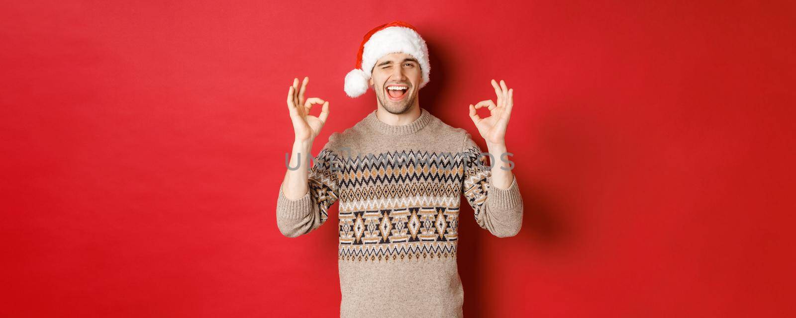 Image of joyful attractive guy in sweater and santa hat, wishing merry christmas, showing okay signs and winking at camera, celebrating new year, standing over red background by Benzoix