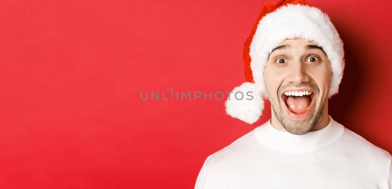 Concept of winter holidays, christmas and celebration. Close-up of surprised handsome man in santa hat, hear amazing new year promo offer, standing against red background.