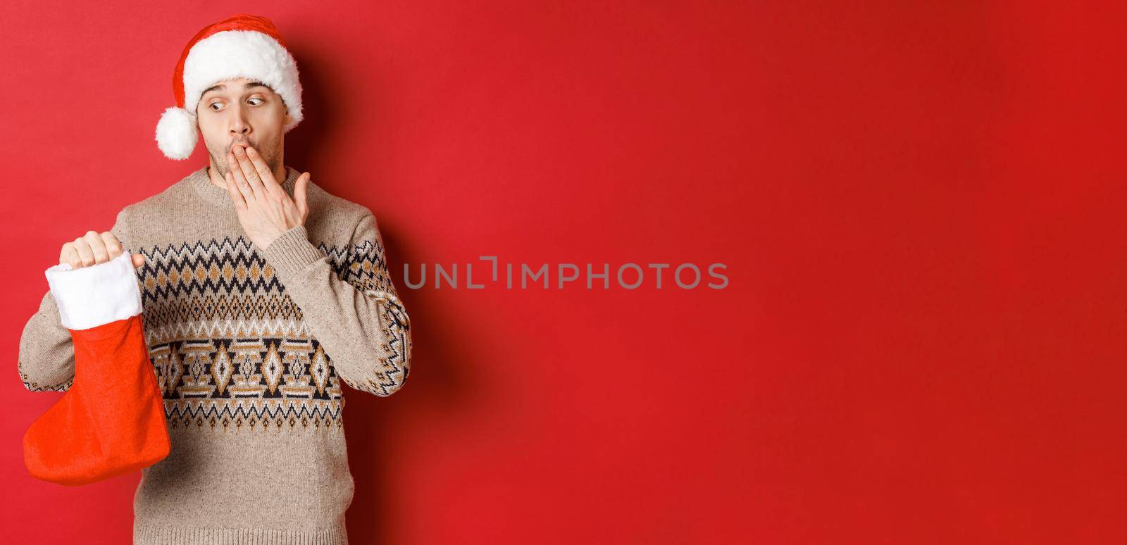 Concept of winter holidays, new year and celebration. Surprised adult man holding christmas stocking with presents inside, looking amazed at bag, standing over red background in santa hat by Benzoix