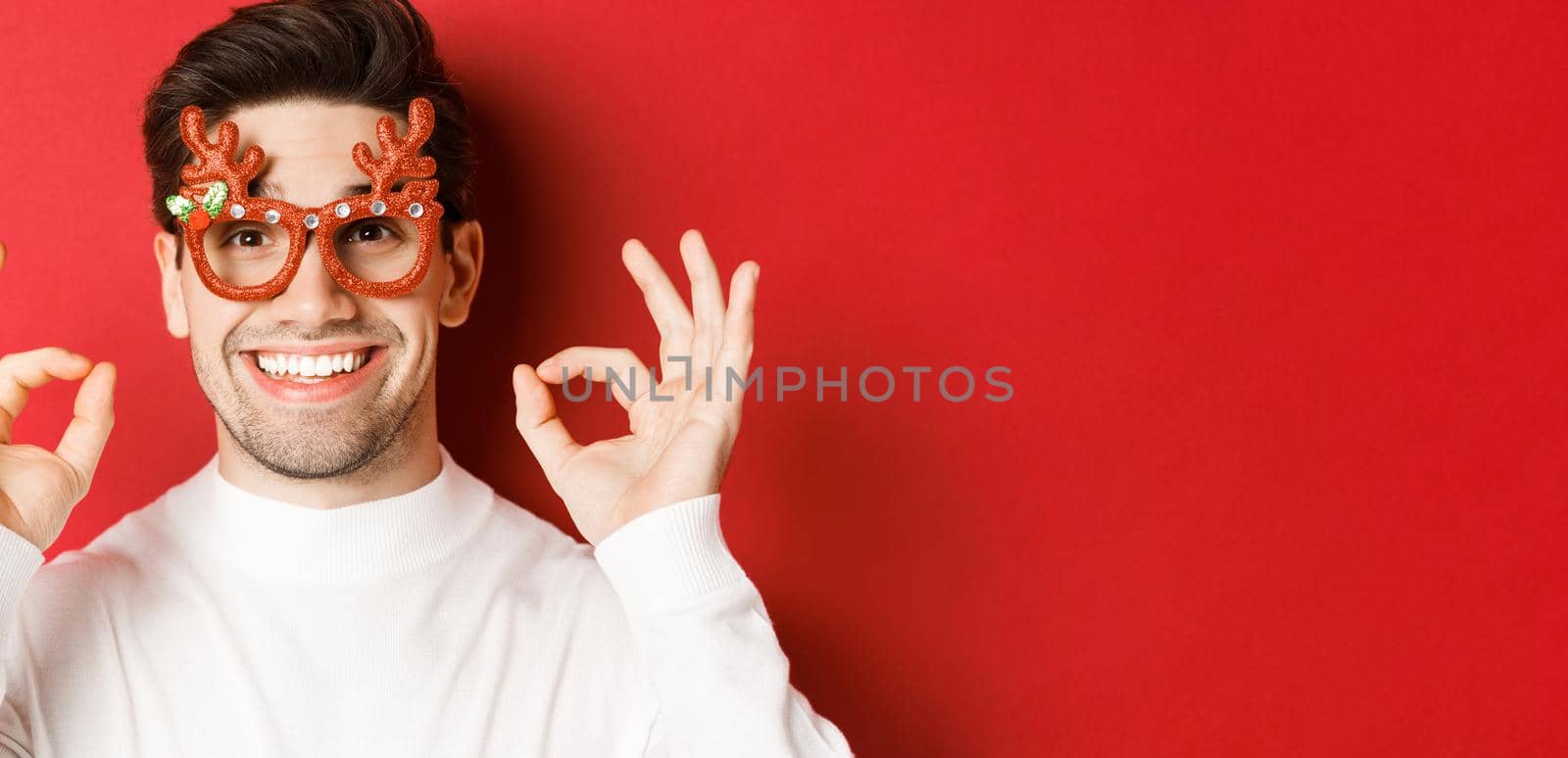 Concept of winter holidays, christmas and celebration. Close-up of attractive man in party glasses, showing okay signs and smiling, praise something good, red background by Benzoix