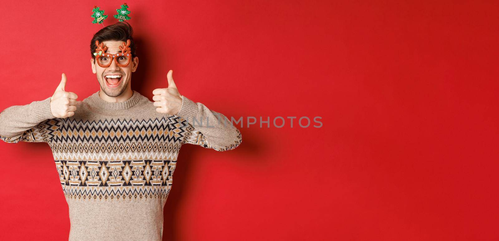 Portrait of satisfied and happy man in christmas sweater and party glasses, showing thumbs-up, wishing happy new year, standing over red background by Benzoix