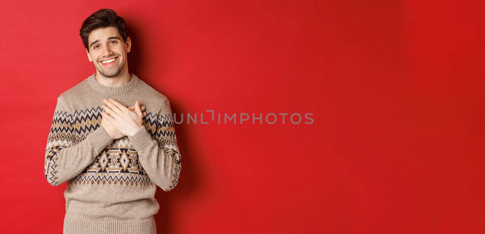 Portrait of touched and happy handsome guy, receiving new year gift, holding hands on heart and smiling, saying thank you, standing in christmas sweater against red background.