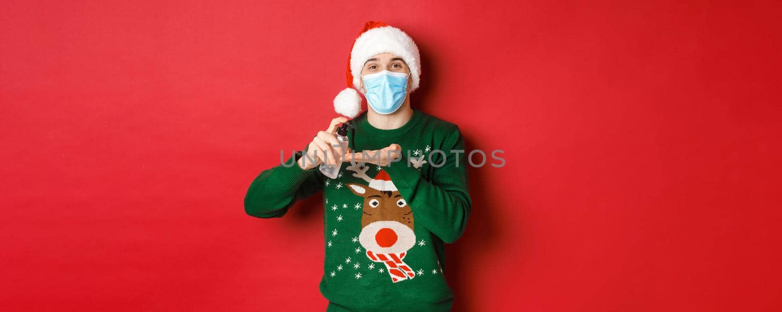 Concept of new year, coronavirus and social distancing. Attractive young man in santa hat, medical mask and christmas sweater, using hand sanitizer to clean hands, standing over red background by Benzoix