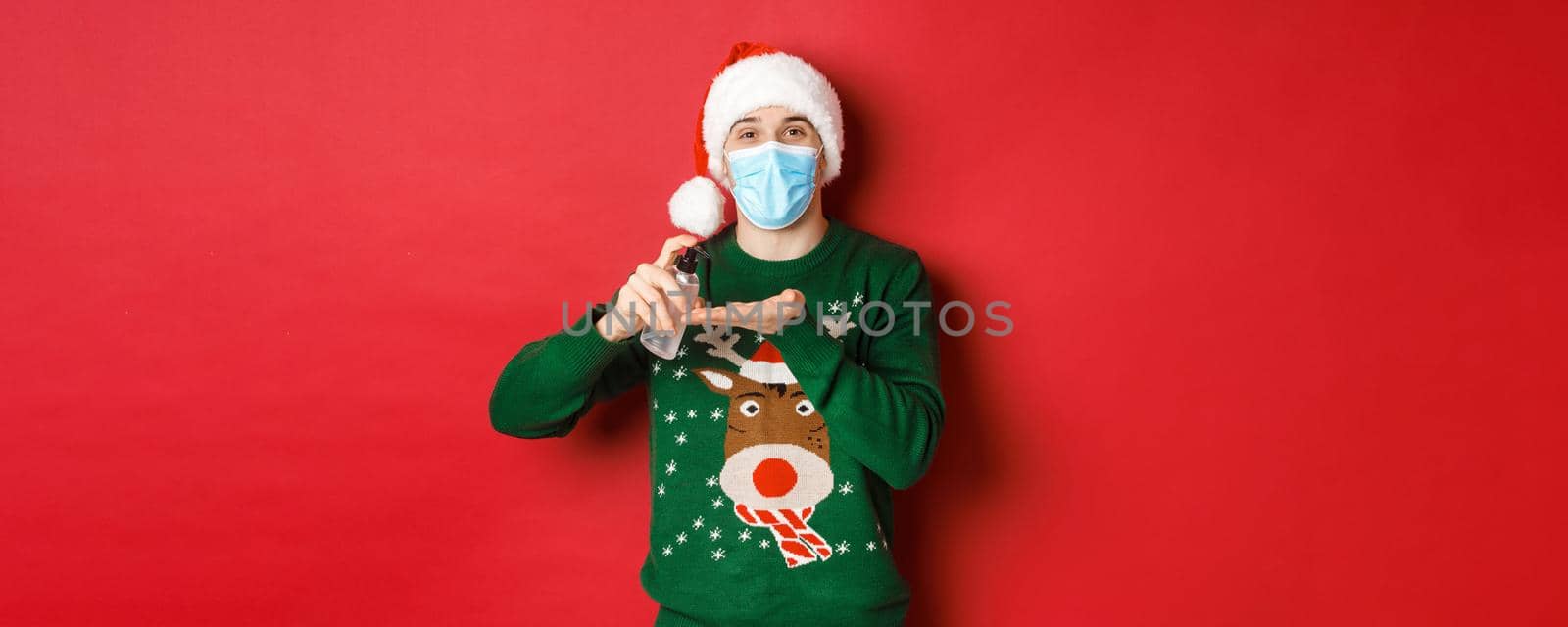 Concept of new year, coronavirus and social distancing. Attractive young man in santa hat, medical mask and christmas sweater, using hand sanitizer to clean hands, standing over red background.