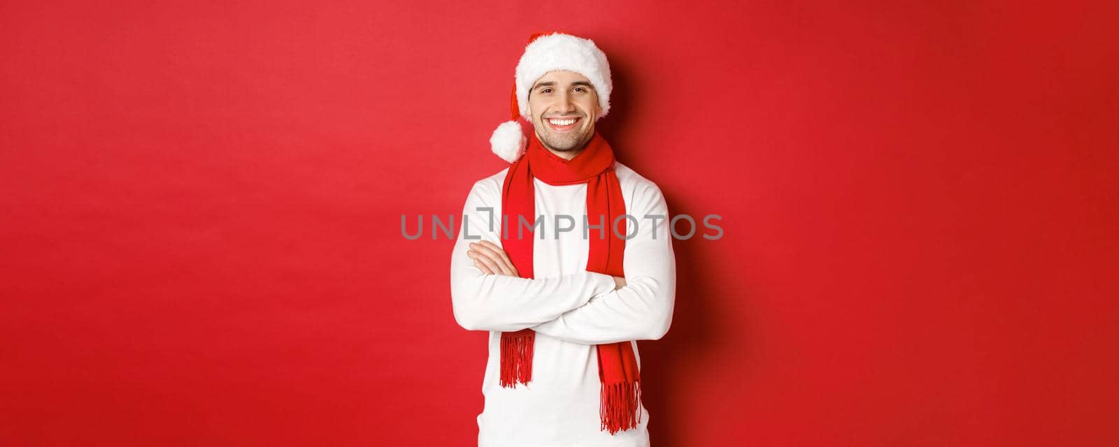 Handsome caucasian guy with bristle, wearing santa hat, scarf and white sweater, cross arms on chest and smiling happy, standing against red background by Benzoix