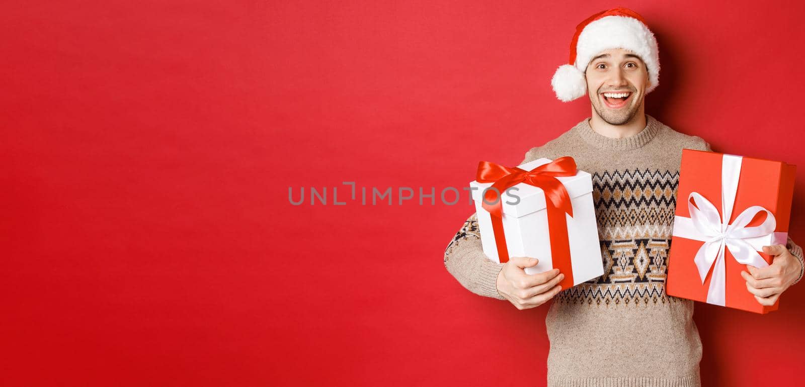 Image of cheerful attractive guy holding christmas presents, standing in santa hat and winter sweater, smiling happy, standing over red background.