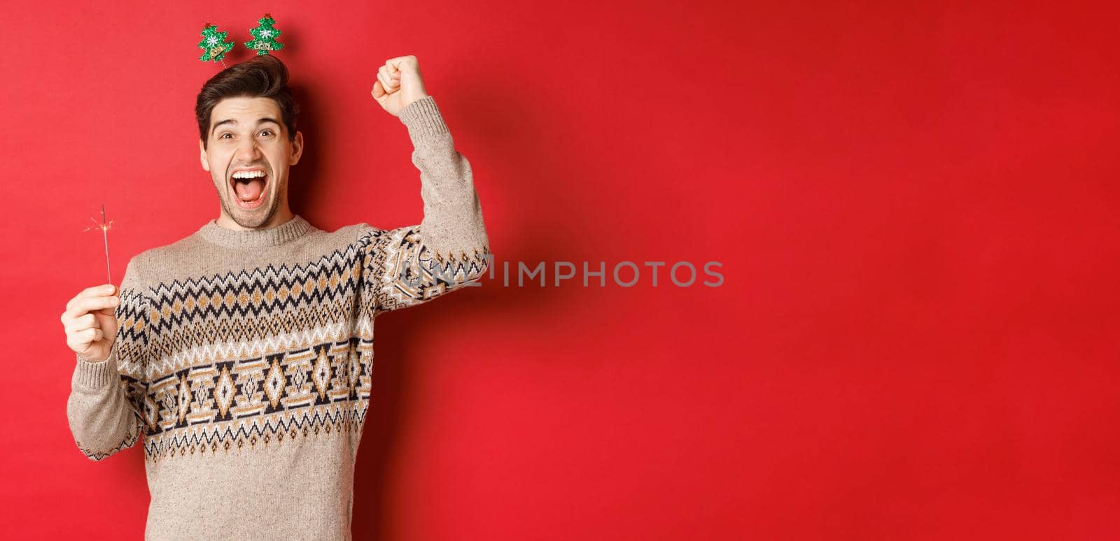 Cheerful guy with a sparkler rejoicing and wishing happy new year, raising hand up and shouting for joy, standing over red background by Benzoix