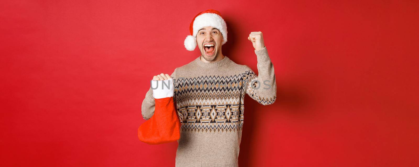 Concept of winter holidays, new year and celebration. Amazed and happy man shouting for joy, found gift inside christmas stocking and cheering, raising hand up and smiling.