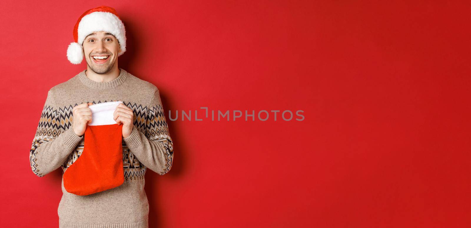 Concept of winter holidays, new year and celebration. Image of surprised and happy man receiving gift from secret santa inside christmas stocking bag, open gift and looking thankful by Benzoix