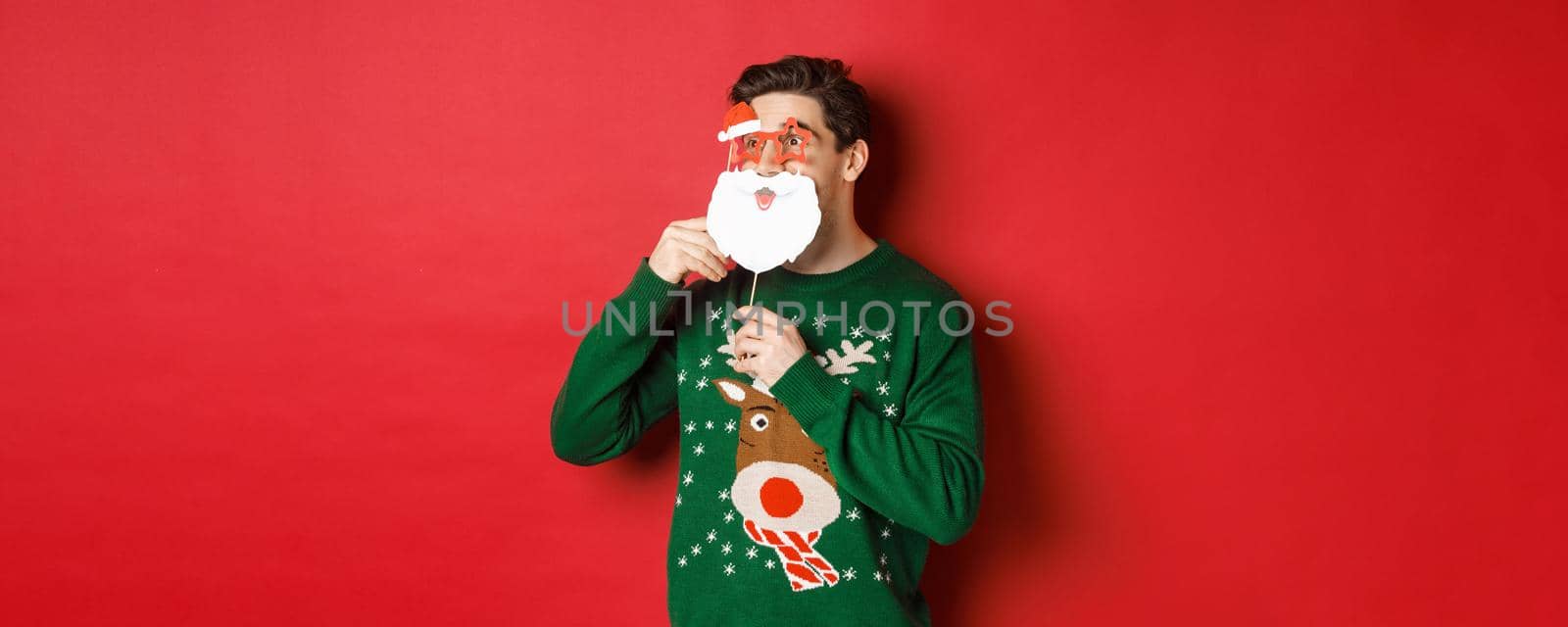 Portrait of surprised man in green christmas sweater, holding funny santa claus mask, celebrating new year, looking left amazed, sanding over red background.