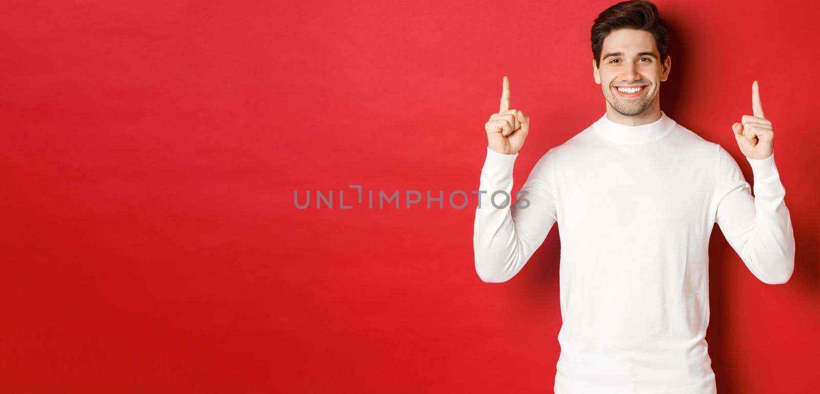 Concept of winter holidays. Good-looking young man with bristle, wearing white sweater, showing christmas advertisement on copy space, pointing fingers up and smiling, red background by Benzoix