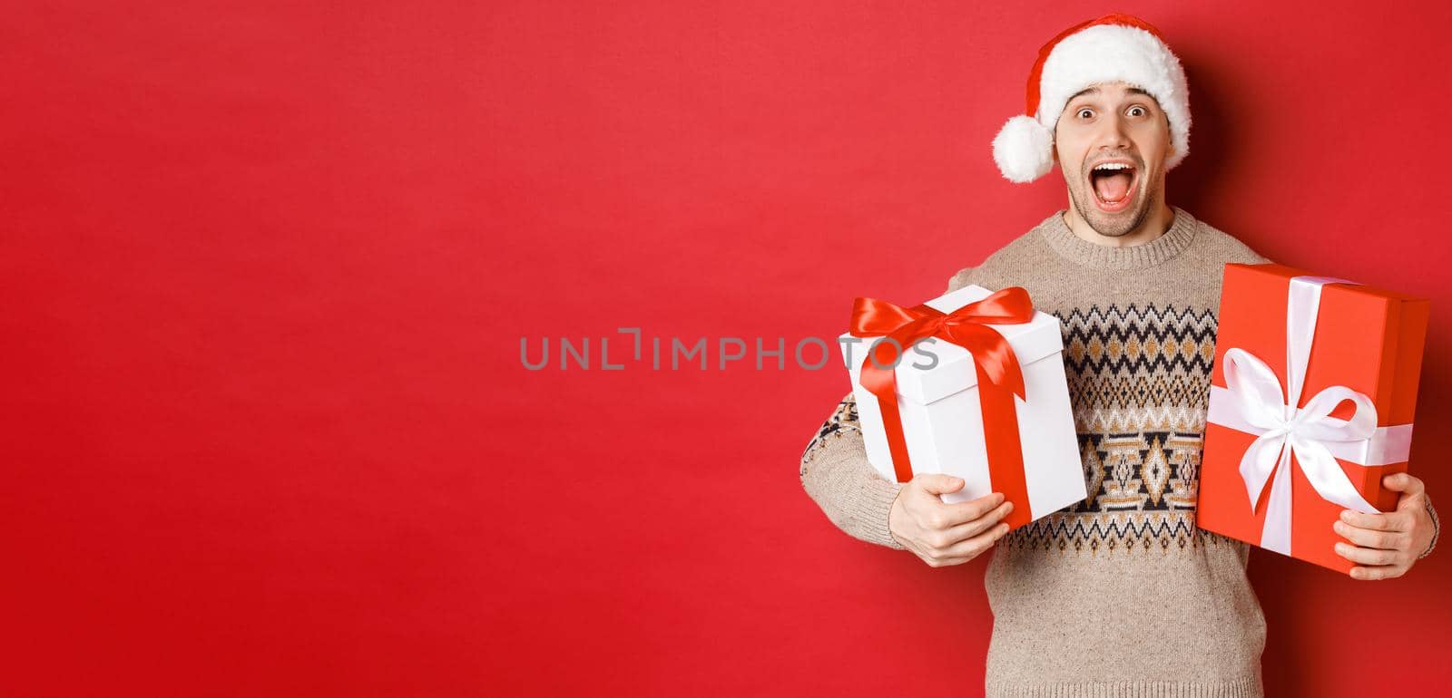 Image of cheerful attractive guy holding christmas presents, standing in santa hat and winter sweater, smiling amazed, standing over red background by Benzoix