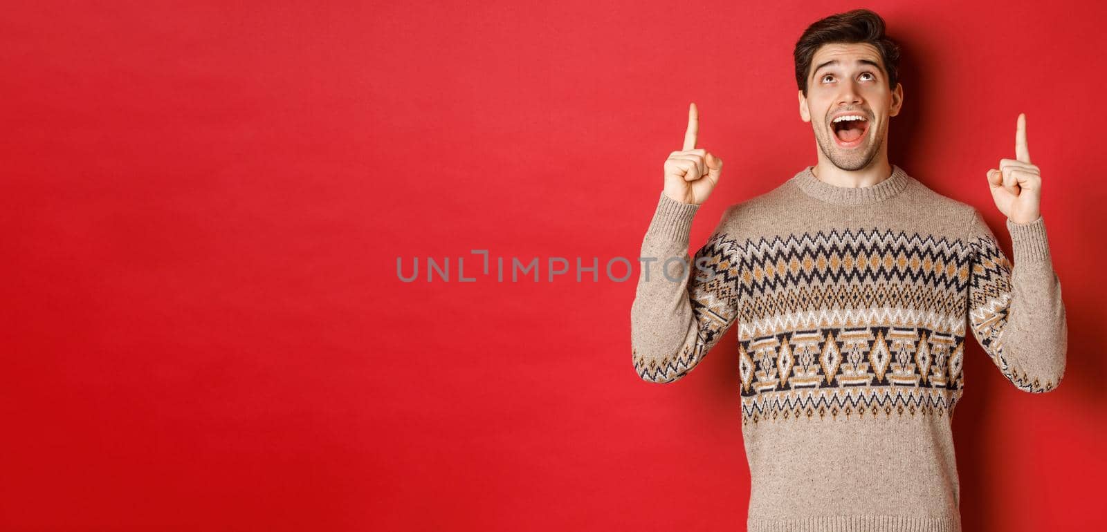 Image of excited handsome guy who likes winter holidays showing advertisement, looking and pointing fingers up with amazed face, standing in christmas sweater over red background by Benzoix