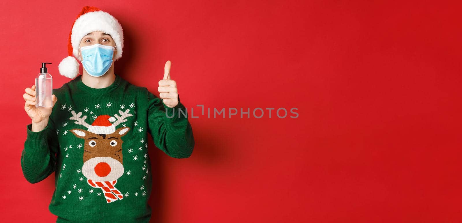 Concept of new year, coronavirus and social distancing. Portrait of cheerful man in santa hat, christmas sweater and medical mask, showing thumb-up and recommending hand sanitizer.