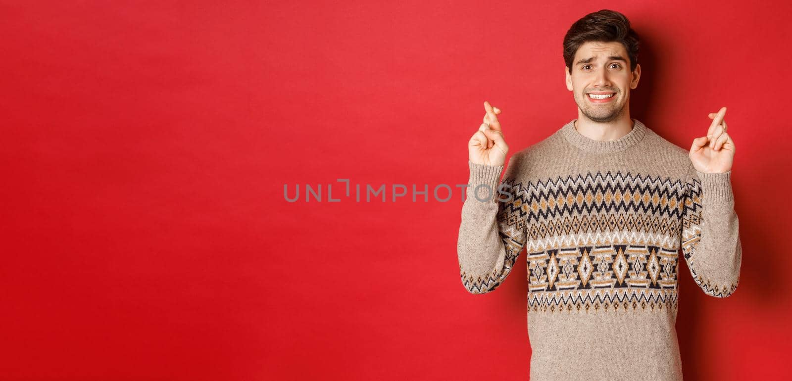 Image of hopeful and worried man in christmas sweater waiting for something, cross fingers for good luck and making wish, nervous about new year gift, red background.