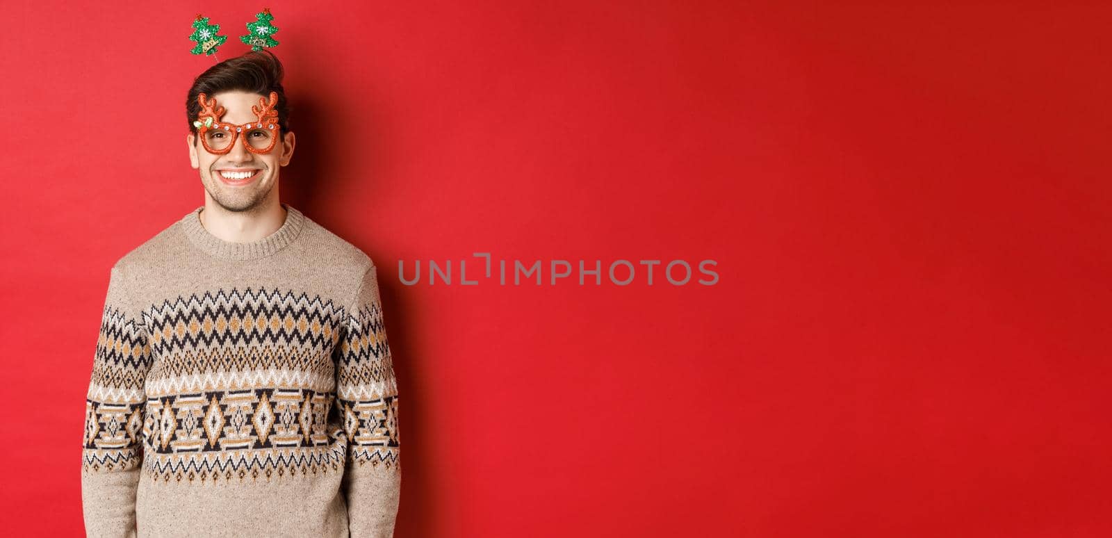 Portrait of handsome bearded guy in party glasses and winter sweater, smiling pleased, celebrating new year, wishing merry christmas, standing over red background by Benzoix