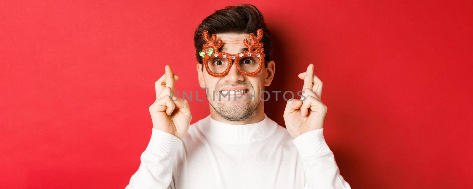 Concept of winter holidays, christmas and celebration. Close-up of nervous man in party glasses, crossing fingers for good luck and pleading, making a wish, standing over red background.