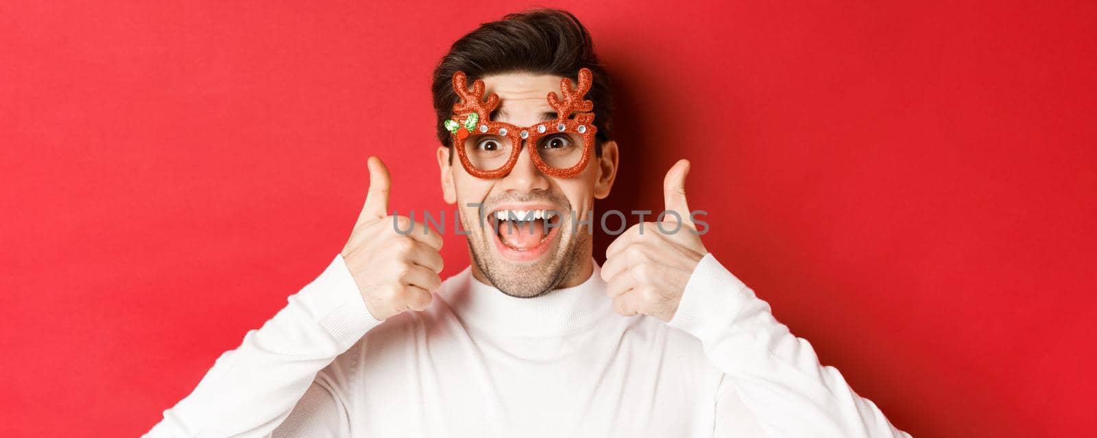 Concept of winter holidays, christmas and celebration. Close-up of excited handsome man in party glasses, smiling and showing thumbs-up in approval, standing over red background by Benzoix