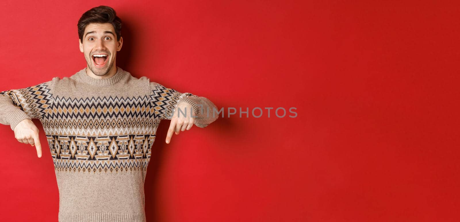Concept of christmas celebration, winter holidays and lifestyle. Excited caucasian male model in xmas sweater, pointing fingers down and making announcement, showing advertisement, red background.